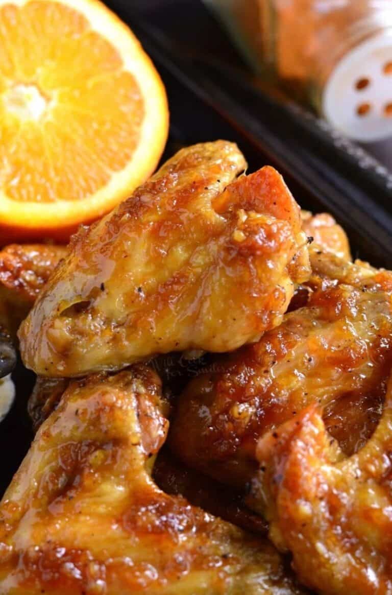 A stack of orange glazed chicken wings on a baking sheet with an orange on the side.